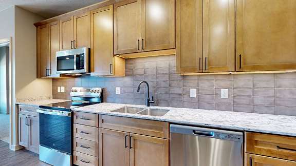 Modern apartment kitchen with wood cabinetry, quartz-style countertops, a tile backsplash, stainless steel appliances, and a double-basin sink beneath under-cabinet lighting.