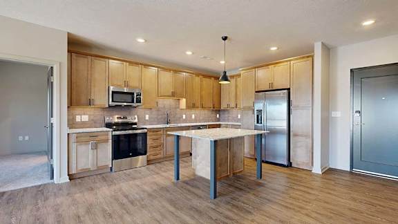 Open-concept apartment kitchen with a center island, wood cabinetry, stainless steel appliances, and wood-look flooring flowing into the adjacent living area.