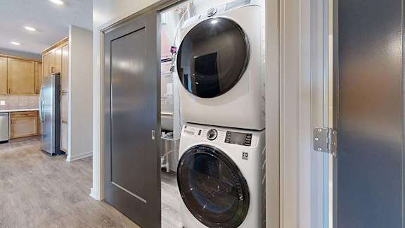 Apartment laundry closet with a stacked front-load washer and dryer, conveniently located off the kitchen with wood-look flooring and modern interior finishes.