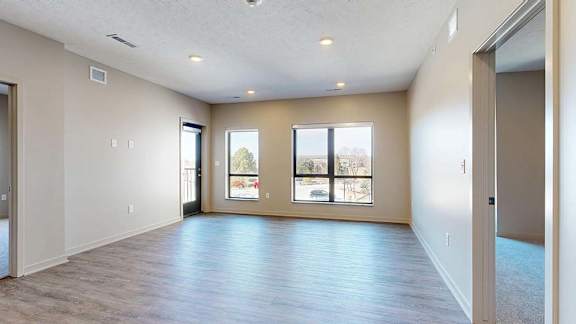 Bright, open apartment living room with wood-look flooring, recessed lighting, large windows, and a sliding glass door that brings in natural light.