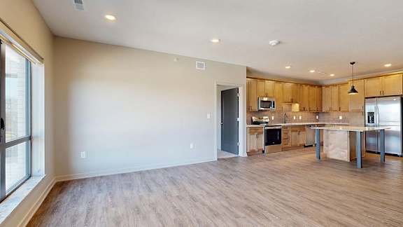 Bright open-concept apartment living room with wood-look flooring, large windows, and a modern kitchen featuring a center island, wood cabinetry, and pendant lighting in the background.
