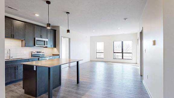 a modern kitchen with a table island and dark cabinets next to a living room with two large windows