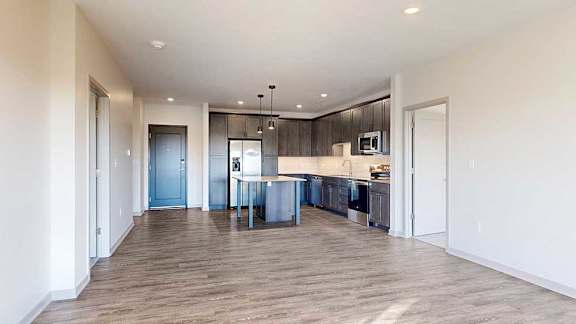 a modern kitchen with a central table island and dark wooden cabinets next to a spacious living room