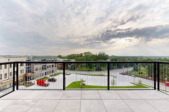 An oversized balcony with a black metal railing, stone tiles, and a view of the neighborhood.