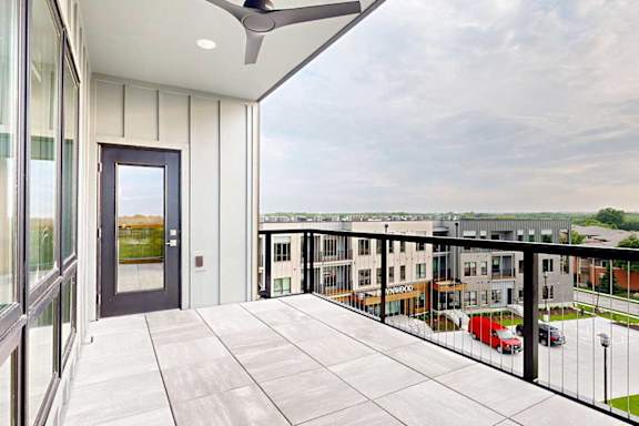 An oversized balcony with a metal and wire railing, stone tile flooring, a ceiling fan, and a windowed door to the living room.