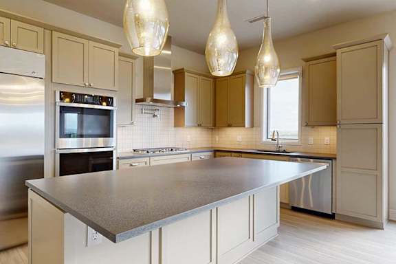 A gourmet kitchen with an oversized island, taupe wooden cabinetry, stainless steel commercial appliances, a range hood, a tile backsplash, and pendant lighting.