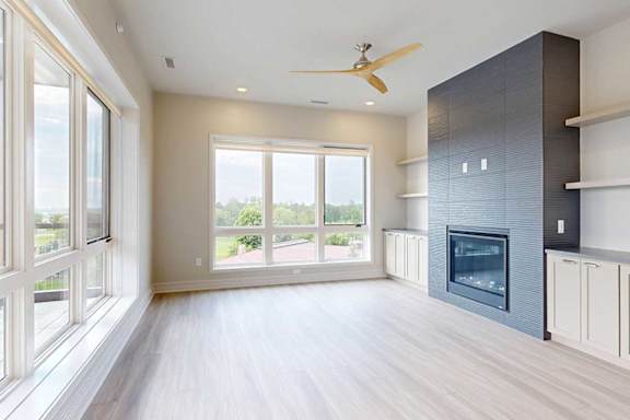 A modern living room with a fire place in a floor-to-ceiling mantel, built-in cabinetry, floor-to-ceiling windows, hardwood-style flooring, and a ceiling fan.