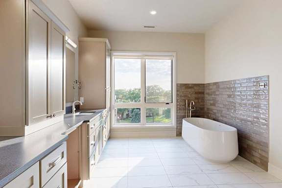 A spacious ensuite bathroom with a double vanity sink, floor-to-ceiling window, soaking tub, and tiled flooring.