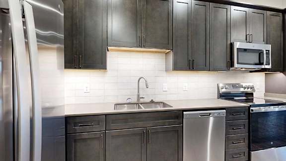 a kitchen with dark wooden cabinets, stainless steel appliances, tile backsplash, and quartz countertops