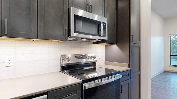 a kitchen with dark wooden cabinets, tile backsplash, quartz countertops, and a stainless steel microwave and oven