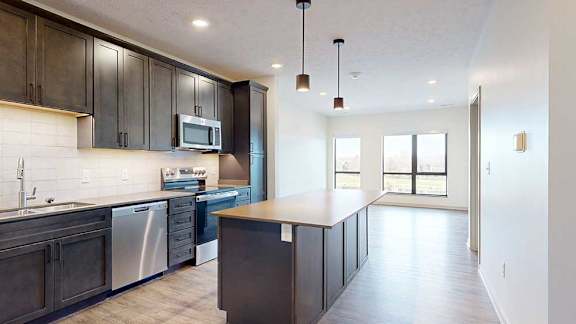 a modern kitchen with dark wood cabinets, light quartz countertops, a large central island, a tile backsplash, and stainless steel appliances next to a living room with large windows