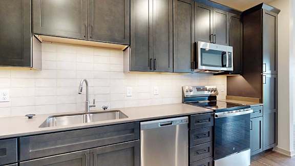 a modern kitchen with dark wood cabinets, light quartz countertops, a tile backsplash, and stainless steel appliances