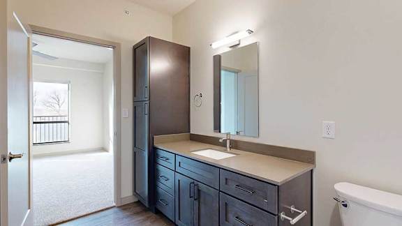 A walk-through bathroom with dark wooden cabinets, a wide quartz vanity, and a door open to a bedroom.