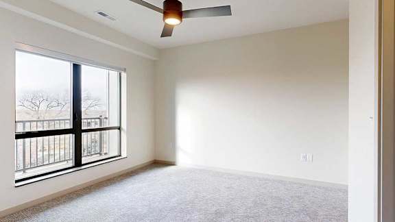 A carpeted bedroom with a ceiling fan and a large window looking out to the balcony.