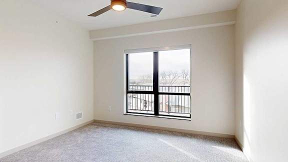 A carpeted bedroom with a ceiling fan and a large window looking out to the balcony.