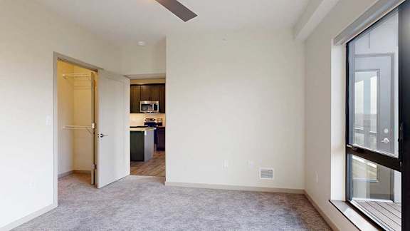 A carpeted bedroom with a floor-to-ceiling window, a walk-in closet, and an open door to a kitchen.