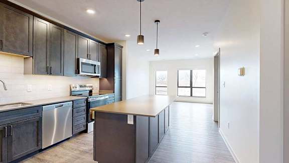 A kitchen with dark brown cabinets and a large island, in front of a living room with floor to ceiling windows.