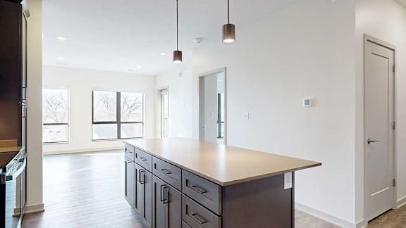 A kitchen with a large table island with cabinets and pendant lights in front of a spacious living room with floor-to-ceiling windows.