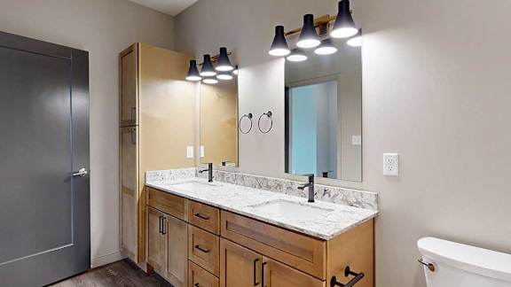 A bathroom with warm wooden cabinets, a quartz double vanity sink, two mirrors with overhead lighting, and tall storage cabinet.