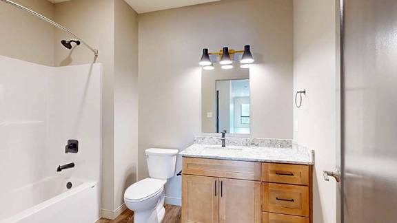 A bathroom with warm wooden cabinets, a white quartz vanity sink, mirror with overhead lighting, and tub shower with black fixtures.