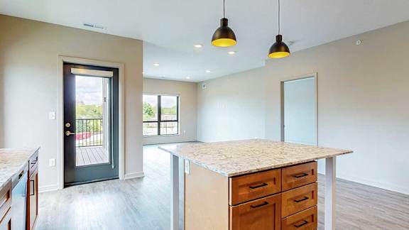 A kitchen with a large table island with cabinets and pendant lights in front of a spacious living room with floor-to-ceiling windows.