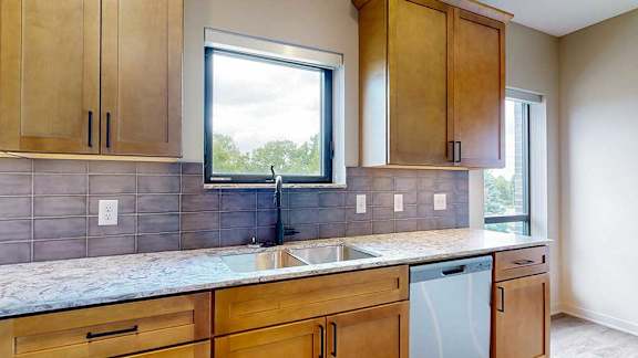 A modern kitchen sink with quartz counters, warm wooden cabinets, a tile backsplash, and a window over the sink.
