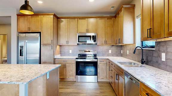 a kitchen with light brown wooden cabinets, stainless steel appliances, a tile backsplash, a large central island, and quartz countertops