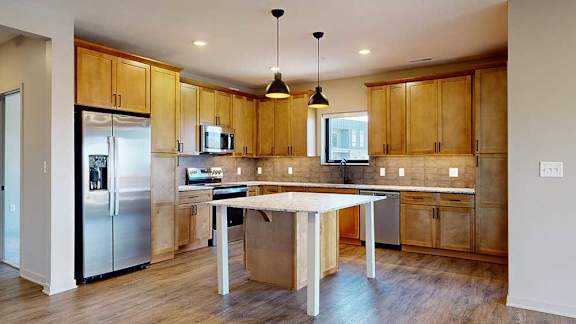 a kitchen with light brown wooden cabinets, stainless steel appliances, a tile backsplash, a large central island, and quartz countertops