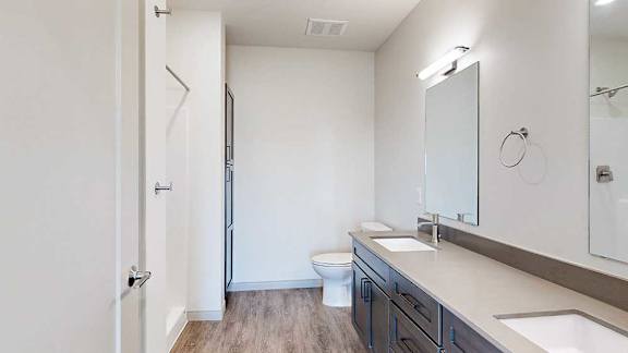 A bathroom with dark wooden cabinets, a quartz double vanity sink, two mirrors with overhead lighting, and a walk-in shower.
