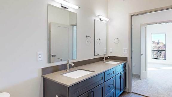 A bathroom with dark wooden cabinets, a quartz double vanity sink, two mirrors with overhead lighting, and a door open to a walk-through closet.