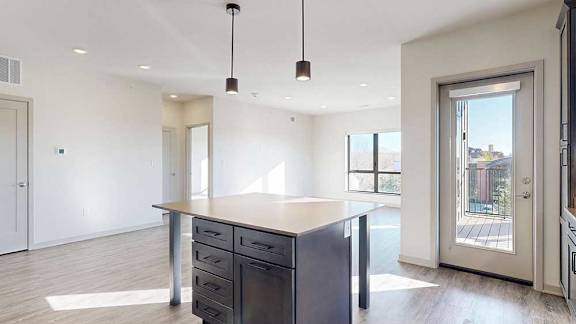 A kitchen with dark wooden cabinets and a large island, in front of a spacious living room with floor-to-ceiling windows.