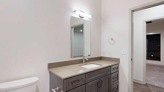 a bathroom with a quartz countertop vanity sink featuring dark wood cabinets, a slim mirror, and overhead lighting