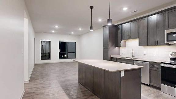 a modern kitchen with dark cabinets next to a long living room with two windows