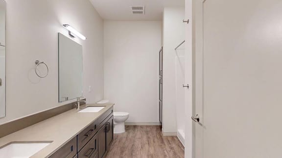 Apartment bathroom with a double-sink vanity, wood-style flooring, and bright lighting.