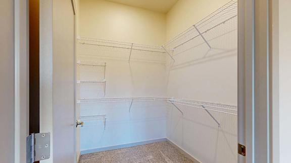 Spacious walk-in closet with white wire shelving on three walls, neutral carpet, and bright overhead lighting in an apartment interior.