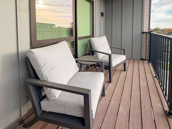 Apartment balcony with wood-style flooring, two cushioned chairs, and a small table beside a large window.
