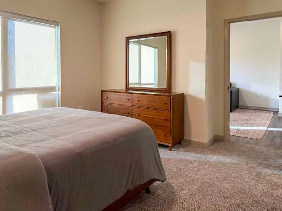 Bright bedroom with carpet flooring, large window, bed, and wood dresser with mirror, opening to an adjacent living area.