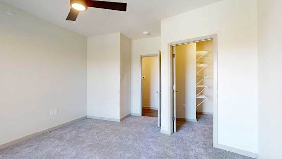 A carpeted bedroom with a walk-in closet, ceiling fan, and a door open to the living room.