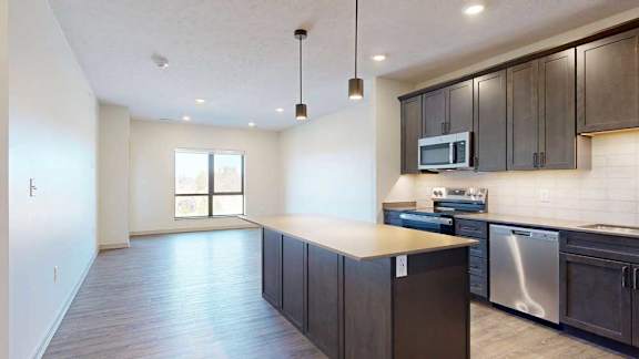 A kitchen with dark wooden cabinets and a large island, in front of a spacious living room with floor-to-ceiling windows.
