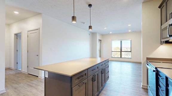 A kitchen with dark wooden cabinets and a large island, in front of a spacious living room with floor-to-ceiling windows.