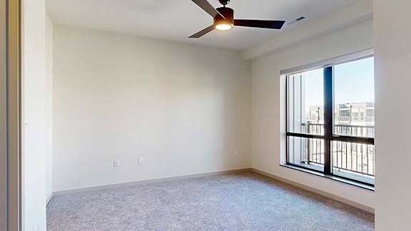 A bedroom with a ceiling fan and an oversized window overlooking a balcony.