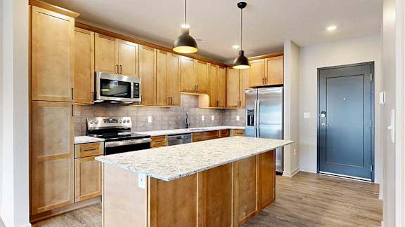 a modern kitchen with wooden cabinets, quartz countertops, a tile backsplash, under cabinet lighting, and stainless steel appliances