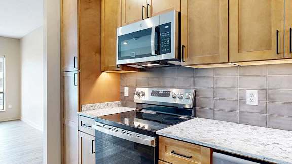 a modern kitchen with wooden cabinets, quartz countertops, a tile backsplash, under cabinet lighting, and stainless steel appliances