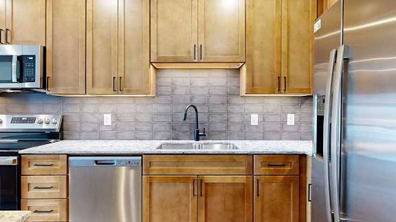 a modern kitchen with wooden cabinets, quartz countertops, a tile backsplash, under cabinet lighting, and stainless steel appliances