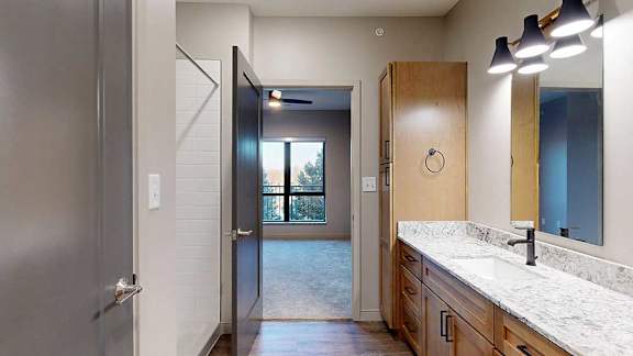 a bathroom with tan wooden cabinets, quartz countertops, a tall storage cabinet, and a door to the bedroom