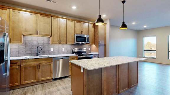 a kitchen with tan wooden cabinets, a large central island, quartz countertops, tile backsplash, and stainless steel appliances