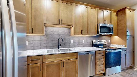 a kitchen with tan wooden cabinets, a tile backsplash, quartz countertops, and stainless steel appliances