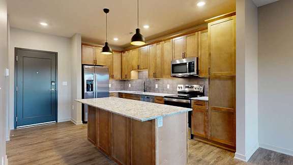a kitchen with tan wooden cabinets, a large central island, quartz countertops, tile backsplash, and stainless steel appliances