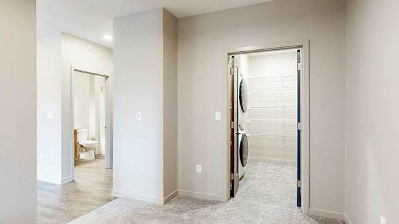 Sleeping area with carpet flooring, view into walk-in closet with stacked washer and dryer, and hallway leading to entry and bathroom.
