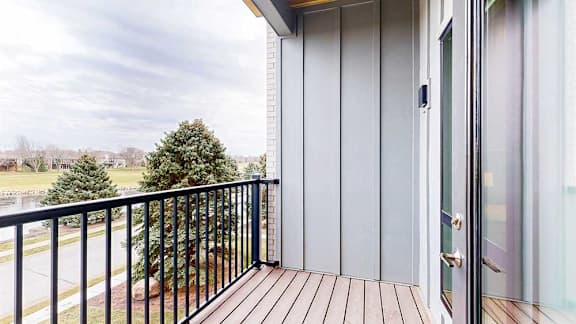 A balcony view with wood-style decking and black metal railing, overlooking greenery.